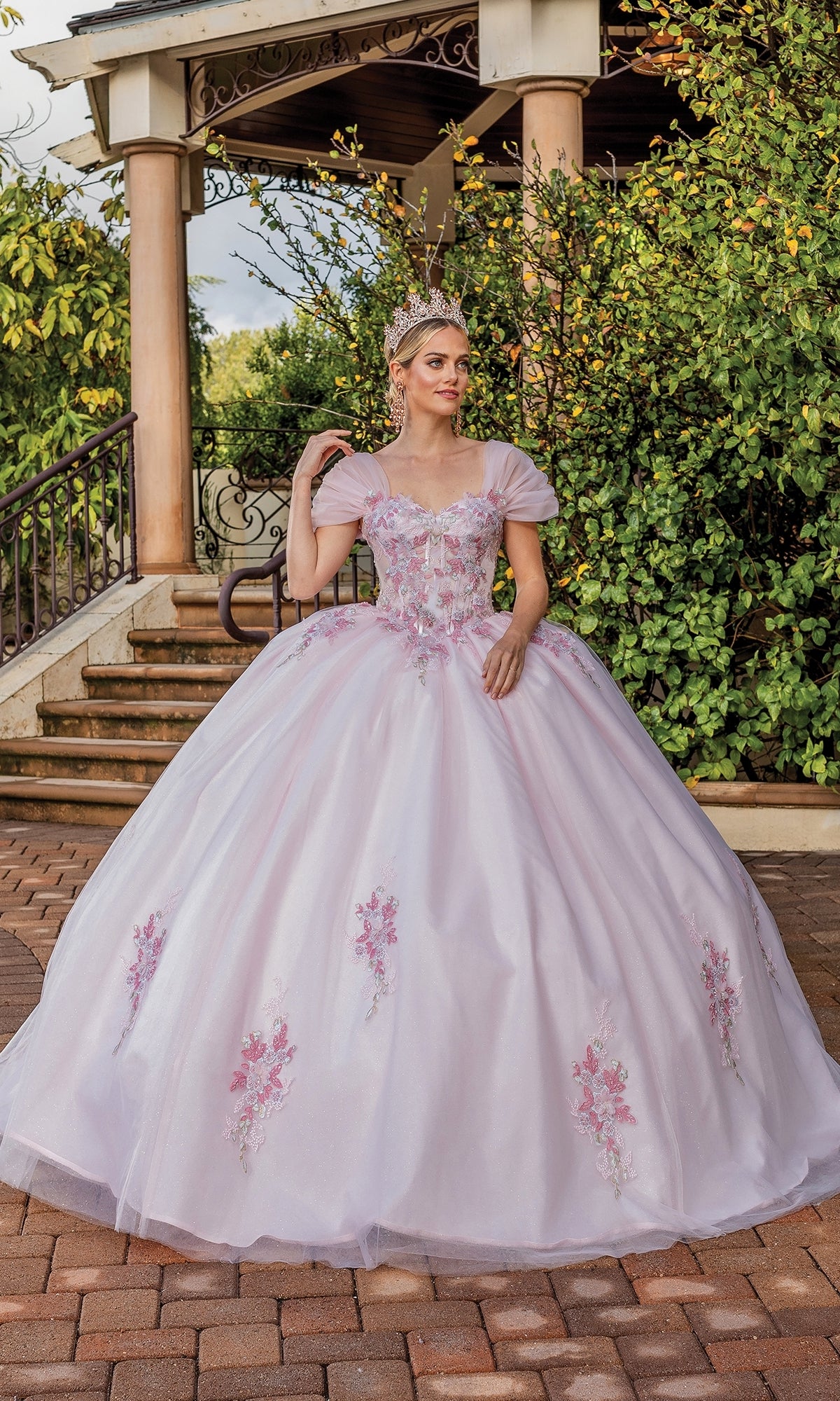 A woman in the Quinceanera Dress 1892 By Dancing Queen, a blush short sleeve a-line gown with floral details and a tiara, stands on a brick path surrounded by greenery and a gazebo.